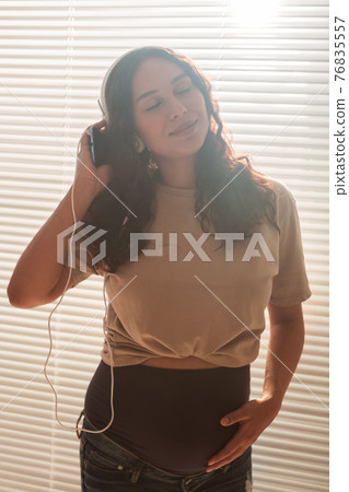 Curly-haired brunette pacified pregnant woman listens to pleasant music using smartphone and headphones. Concept of a soothing mood before meeting baby. 76835557