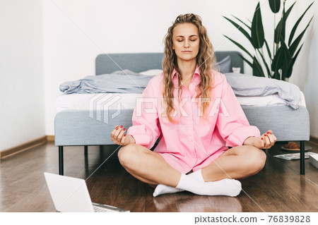 A young woman meditates while sitting next to a bed in . The girl does yoga exercises at home to relieve stress, taking a break from work. A young woman meditates while sitting next to a bed in . The girl does yoga exercises at home to relieve stress, taking a break from work. 76839828