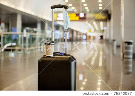 In an empty airport, there is a suitcase, a glass of coffee on it, and a medical protective mask. Coronavirus, social distance, isolation 76840087