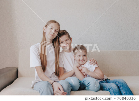 three children, a girl and two boys in white T-shirts and blue jeans, sit hugging each other on the home sofa and smile. 76840336