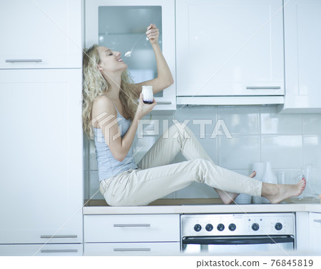 Side view of young woman eating yogurt while sitting on kitchen counter 76845819