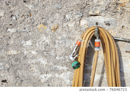 Coiled garden hose hanging on faucet against stone wall 76846723