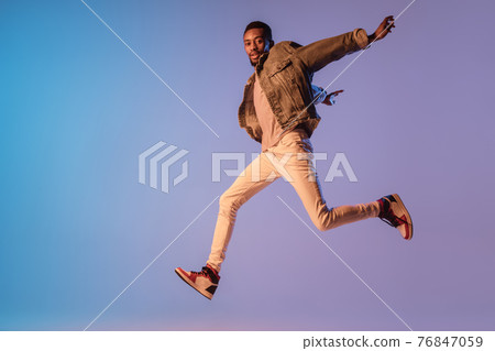 Young stylish man in modern street style outfit isolated on gradient background in neon light. African-american fashionable model in look book, musician performing. 76847059