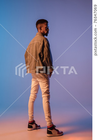 Young stylish man in modern street style outfit isolated on gradient background in neon light. African-american fashionable model in look book, musician performing. Young stylish man in modern street style outfit isolated on gradient background in neon light. African-american fashionable model in look book, musician performing. 76847069