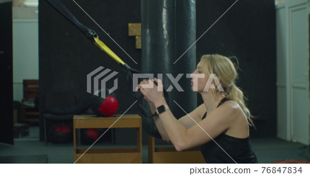 Blonde woman in sportswear doing exercise with TRX system at gym. Cross training. Modern fitness. Blonde woman in sportswear doing exercise with TRX system at gym. Cross training. Modern fitness. 76847834