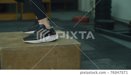 Close up young female with perfect body doing set of box jumps at the gym. Fitness woman in sportswear doing jumps on a big box in a training centre. 76847871