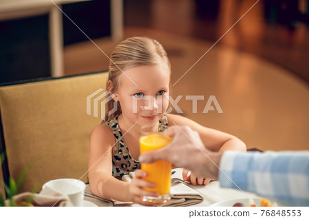Cute girl sitting at the table and drinking orange juice Cute girl sitting at the table and drinking orange juice 76848553