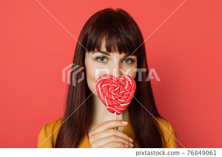 Close up studio shot of charming young red haired woman, wearing yellow shirt, holding a heart shaped red sweet lollipop, covering her nose and lips, posing on a red background 76849061