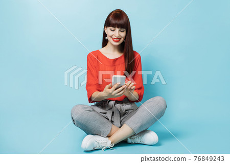 Internet, social networks. Pretty young woman, wearing gray pants and red shirt, sitting on the floor in lotus pose, holds mobile phone, browsing through page of social media isolated on blue. Internet, social networks. Pretty young woman, wearing gray pants and red shirt, sitting on the floor in lotus pose, holds mobile phone, browsing through page of social media isolated on blue. 76849243