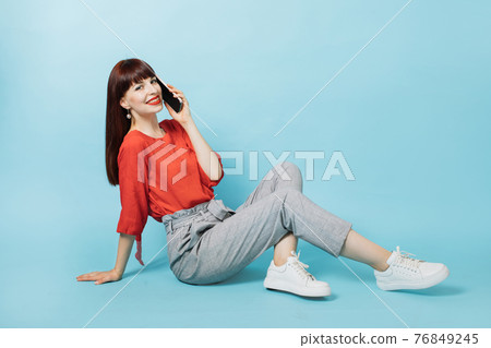 Positive human emotions, mobile communication. Young joyful smiling woman in red shirt and gray pants, bright make up and red lipstick, talking phone, sitting on isolated blue background. Positive human emotions, mobile communication. Young joyful smiling woman in red shirt and gray pants, bright make up and red lipstick, talking phone, sitting on isolated blue background. 76849245