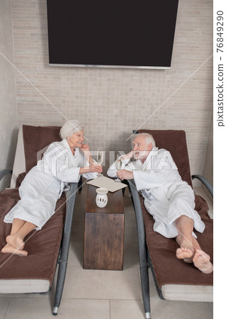 Mature couple in white robes lying on a chaise lounges in a spa center 76849290