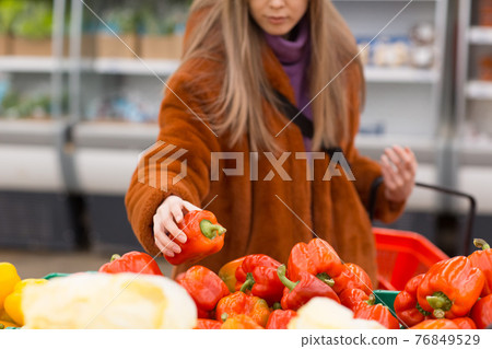 Young woman with a grocery basket chooses red pepper in the vegetable department. Young woman with a grocery basket chooses red pepper in the vegetable department. 76849529