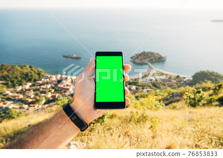 Man's hand holds a smartphone with a blank screen vertically on the background of Sveti Stefan island Montenegro Man's hand holds a smartphone with a blank screen vertically on the background of Sveti Stefan island Montenegro 76853833