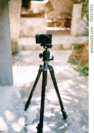 Black digital camera sits on a tripod on the sidewalk. Front view Black digital camera sits on a tripod on the sidewalk. Front view 76853854