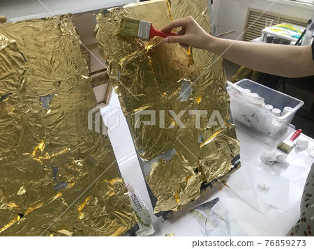The artist's hand paints with a brush using gold glittering bright yellow metallic gold leaf for artistic purposes and creativity on canvas. The background 76859273