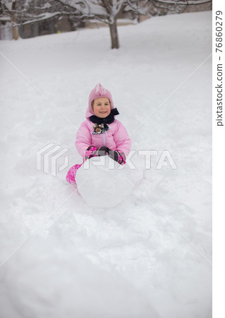 The child plays with snow in the winter. A little girl in a bright jacket and knitted hat, catches snowflakes in a winter park for Christmas. 76860279
