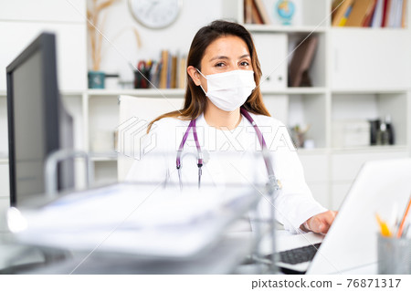 Portrait of a young female doctor in a protective mask working at a computer in the office 76871317