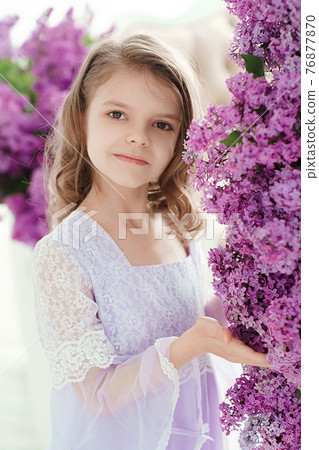 Beautiful little girl of preschool age in a delicate dress sitting on a swing decorated with flowers of lilac. Beautiful little girl of preschool age in a delicate dress sitting on a swing decorated with flowers of lilac. 76877870