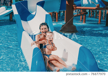Little girl in swimsuit goes down the blue slides to the pool, Mom and daughter play and swim in the outdoor pool Little girl in swimsuit goes down the blue slides to the pool, Mom and daughter play and swim in the outdoor pool 76877970