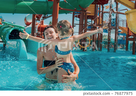 Little girl in swimsuit girl hugs joyful mom in the water park pool. Mother and daughter play and swim in the outdoor pool 76877975