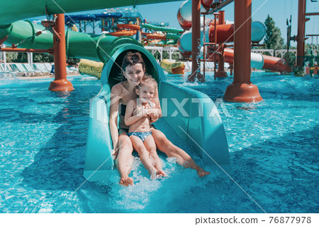 Little girl in swimsuit goes down the blue slides to the pool, Mom and daughter play and swim in the outdoor pool 76877978