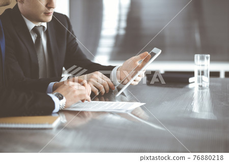 Unknown businessmen using tablet computer and work together at the glass desk in modern office, close-up. Teamwork and partnership concept 76880218