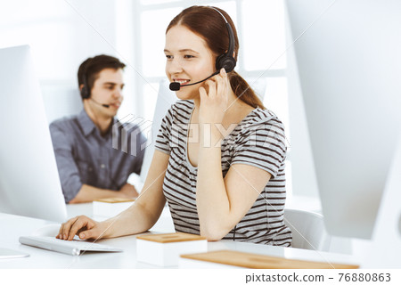 Casual dressed young woman using headset and computer while talking with customers online. Group of operators at work. Call center, business concept Casual dressed young woman using headset and computer while talking with customers online. Group of operators at work. Call center, business concept 76880863
