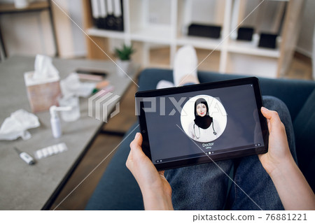 Tablet screen view, close up. Top view of unrecognizable female patient, sitting with digital tablet pc on couch and making video call to young professional Arabian doctor from home Tablet screen view, close up. Top view of unrecognizable female patient, sitting with digital tablet pc on couch and making video call to young professional Arabian doctor from home 76881221