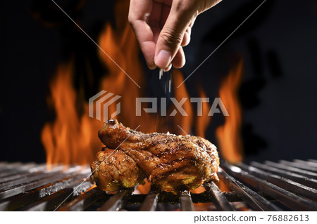 Hand sprinkling salt and spices on grilled chicken leg on the flaming grill 76882613