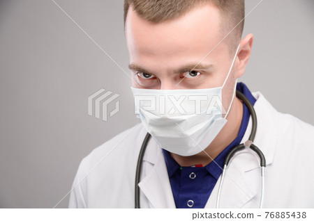 A young Caucasian doctor wearing a mask looks ominously into the camera from under his brow. Medical conspiracy concept against humanity 76885438