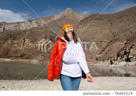 A portrait of a traveler. Women hiking woman happy and smiling, pulling her cap over her eyes and throwing her head back laughs while hiking in the mountains. A portrait of a traveler. Women hiking woman happy and smiling, pulling her cap over her eyes and throwing her head back laughs while hiking in the mountains. 76885524