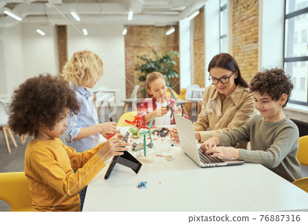 Group of diverse kids working together with young female teacher, using digital devices and playing with mechanical toys while sitting at the table during STEM class 76887316