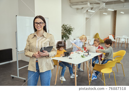 Cheerful young female teacher in casual wear smiling at camera and holding tablet pc while standing in a classroom. Kids working on creating robots in the background 76887318