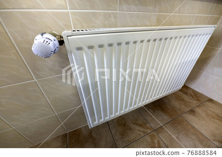 Close-up of white heating radiator with thermostat valve ion tiled walls and floor background. Comfortable warm home interior, climate control, money saving concept. 76888584