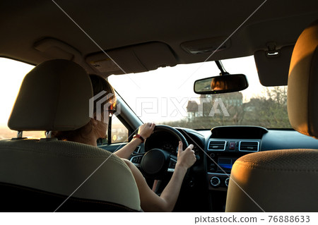 Close up view of a woman driver holding steering wheel driving a car at sunset. Close up view of a woman driver holding steering wheel driving a car at sunset. 76888633