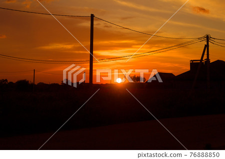 Rural landscape at sunset. Setting sun, houses, road and electrical line on bright orange sky background. Rural landscape at sunset. Setting sun, houses, road and electrical line on bright orange sky background. 76888850