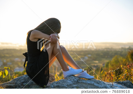 Young depressed woman in black short summer dress sitting on a mountain hill thinking outdoors at sunset. Lonely female contemplating in warm evening in nature. 76889018