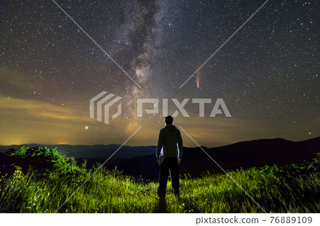 Dark silhouette of a man standing in mountains at night enjoying milky way and Neowise comet with light tail in dark sky view. Dark silhouette of a man standing in mountains at night enjoying milky way and Neowise comet with light tail in dark sky view. 76889109