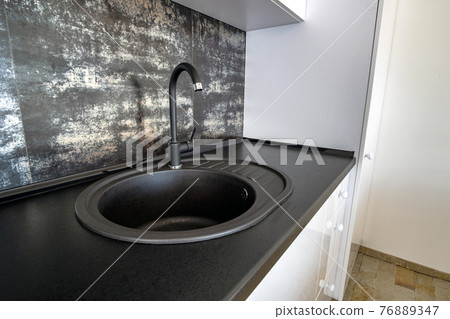Interior of modern spacious kitchen with white contemporary furniture, black ceramic tiles on the wall and dark granite sink with water tap. 76889347