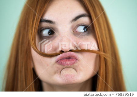 Close up portrait of funny girl holding long red hair like a mustache under her nose. Close up portrait of funny girl holding long red hair like a mustache under her nose. 76889519