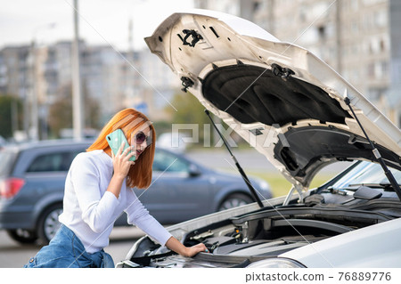 Young woman standing near broken car with popped hood talking on her mobile phone while waiting for help. Young woman standing near broken car with popped hood talking on her mobile phone while waiting for help. 76889776