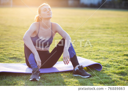 Young sportive woman in sports clothes sitting on training mat before doing exercises in field at sunrise. Young sportive woman in sports clothes sitting on training mat before doing exercises in field at sunrise. 76889863