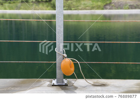 Yellow plastic buoy attached to a metal fence on the bank of a lake with green water. Yellow plastic buoy attached to a metal fence on the bank of a lake with green water. 76889877