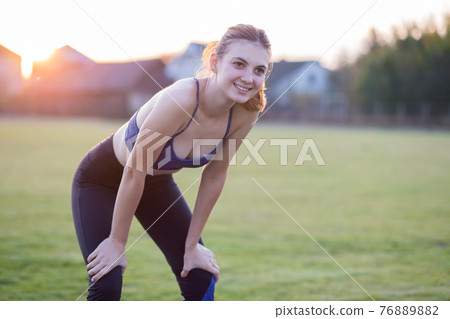 Slim blonde girl plays sports and performs yoga poses in summer grass covered stadium on a sunset background. Woman doing exercises on the yoga mat. 76889882
