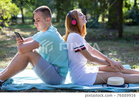 Young teenage couple having fun outdoors in summer park. Girl with red hair listening to music in pink earphones and boy chatting on the sell phone. Young teenage couple having fun outdoors in summer park. Girl with red hair listening to music in pink earphones and boy chatting on the sell phone. 76889916