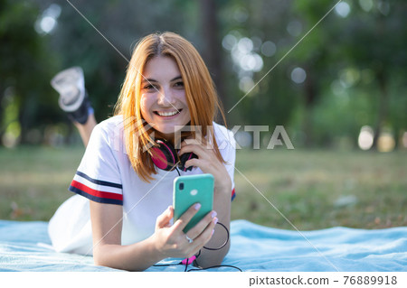 Pretty smiling teenage girl with red hair using sellphone outdoors in park. 76889918