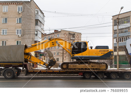 A heavy modern excavator stands on a low trailer platform along the rear ramp. A heavy modern excavator stands on a low trailer platform along the rear ramp. 76889935