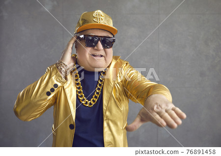 Funny senior man in golden jacket, baseball cap and chain necklace playing music at disco party 76891458