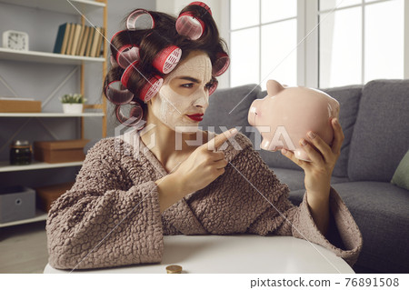 Funny woman in hair curlers shaking finger at piggy bank determined to save up money Funny woman in hair curlers shaking finger at piggy bank determined to save up money 76891508