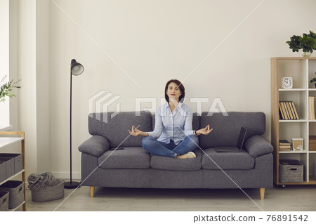 Woman with eyes closed relaxing and meditating in lotus pose on sofa couch Woman with eyes closed relaxing and meditating in lotus pose on sofa couch 76891542